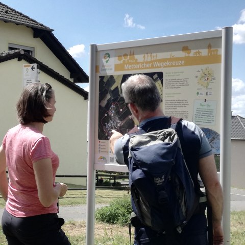 Deux randonneurs se tiennent devant un panneau de randonnée portant l'inscription 'Mettericher Wegekreuze' et s'informent sur l'itinéraire. Des maisons sont visibles à l'arrière-plan., © Tourist-Information Bitburger Land