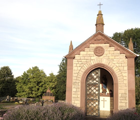 Eine kleine Kapelle in Idenheim mit offenem Eingang, einer Statue im Inneren und umgeben von blühenden Pflanzen und Bäumen., © Thomas Neises