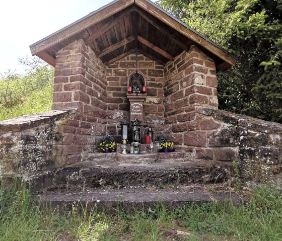 Ein steinernes Wegekreuz mit Dach, umgeben von Pflanzen. Vor dem Kreuz stehen Blumen und Kerzen., © Melanie Lonien