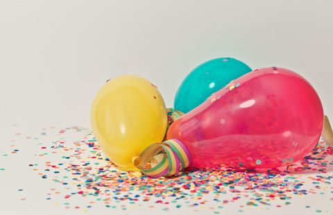 Bunte Luftballons in Gelb, Blau und Pink liegen zusammen auf einem weißen Hintergrund. Rundherum sind funkelnde Konfetti-Stücke verstreut.