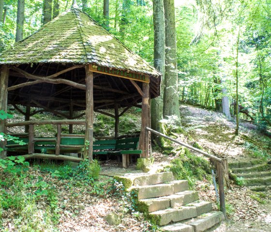 Die Willi Poss Hütte im Wald, umgeben von Bäumen, mit einem moosbedeckten Dach und steinernen Treppenstufen., © TI Bitburger Land_M.Mayer