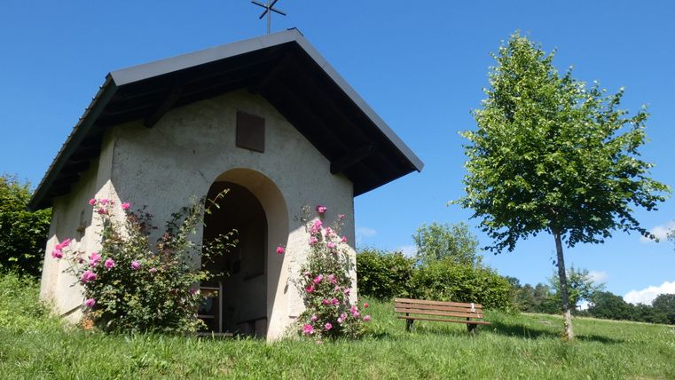 Kleine Kapelle im Grünen mit blühenden Rosen und Bank daneben.