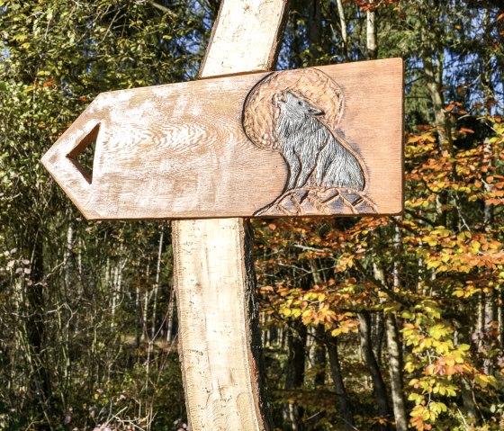 Ein Holzschild mit einem geschnitzten Wolfssymbol zeigt den Weg im herbstlichen Wald. Bunte Blätter im Hintergrund., © TI Bitburger Land