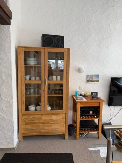 Ein Holzschrank mit Glasfront, der Tassen und Geschirr zeigt. Daneben steht ein kleiner Tisch mit einem Audio-System.