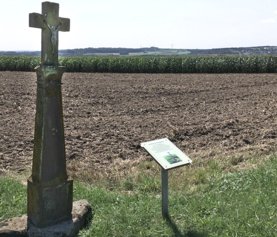 Stone cross with a figure of Jesus in a field near Etteldorf, with an information board next to it. Fields and hills can be seen in the background., © Tourist-Info Bitburger Land M.Mayer
