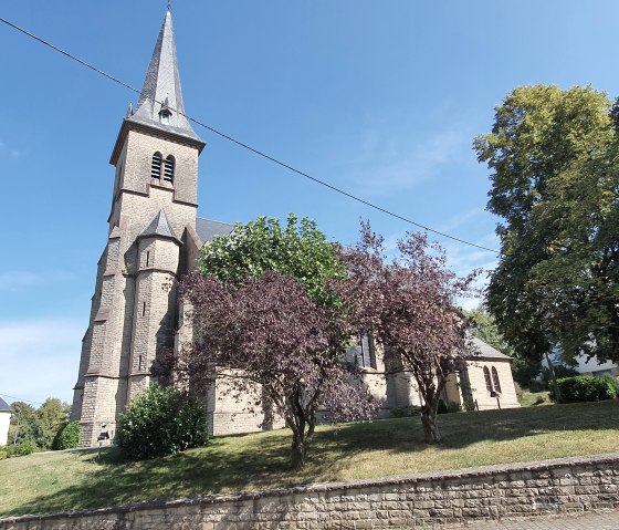 De parochiekerk van St Bartholomew met zijn hoge, spitse toren staat omringd door bomen onder een helderblauwe hemel., &copy; TI Bitburger Land - Steffi Wagner
