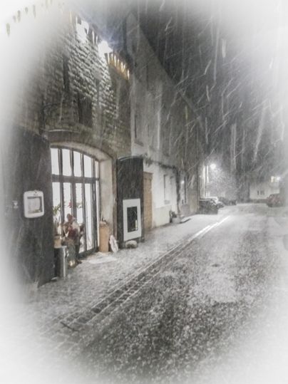 Eine schneebedeckte Straße mit historischen Gebäuden im Hintergrund. Der Schnee fällt sanft in der Nacht und sorgt für eine romantische Atmosphäre.
