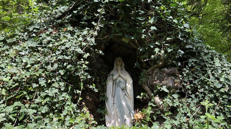 Eine Statue von Maria steht in einer kleinen Höhle, umgeben von grünem Efeu. Die Szene ist idyllisch und natürlich gestaltet.