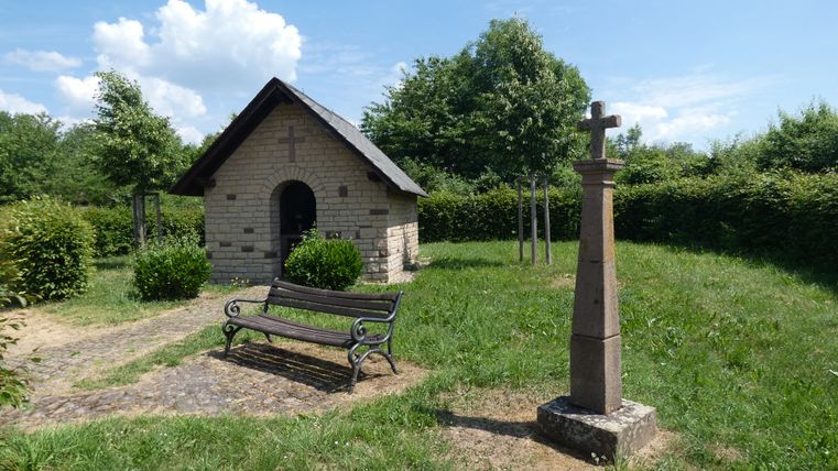 Kleine Kapelle mit Kreuz und Bank in grüner Landschaft.