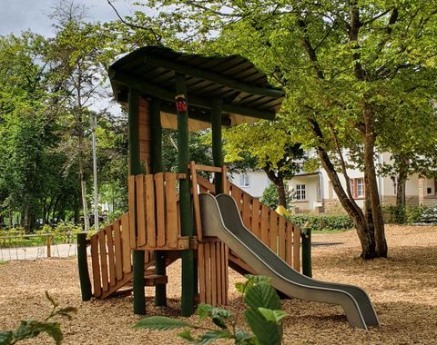 Ein Spielplatz mit einer Holzspielstruktur und einer Rutsche. Der Bereich ist von Bäumen umgeben und hat eine gemütliche Atmosphäre.