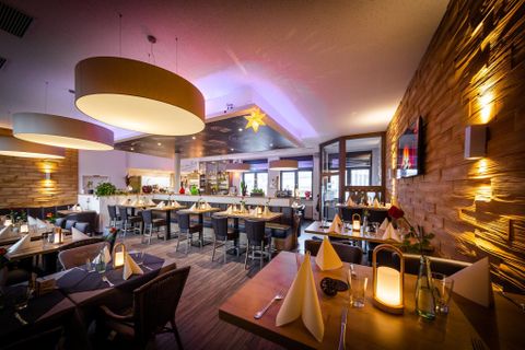 Ein modernes Restaurant mit warmem Licht und elegant eingedeckten Tischen. Die Wände sind mit Holz verziert und die Atmosphäre ist einladend und gemütlich.
