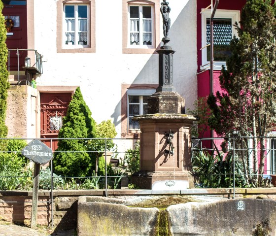 A historic fountain from 1859 stands in front of a building with red and white facades in Dudeldorf. Surrounded by plants and a railing., &copy; Tourist-Information Bitburger Land_M. Mayer