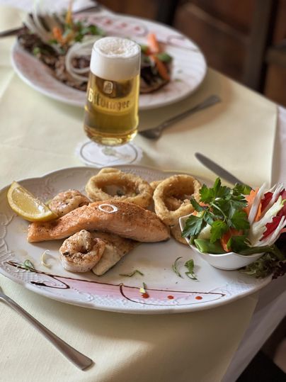 Ein Teller mit gegrilltem Fisch, Garnelen und Calamari, garniert mit Zitronenscheiben und frischem Gemüse. Im Hintergrund steht ein Glas Bier.
