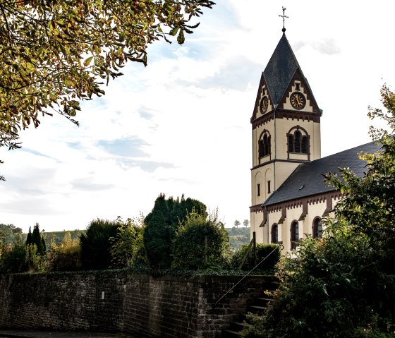 Die Kirche St. Remigius in Oberweis, umgeben von Bäumen und einer Steinmauer, unter einem bewölkten Himmel., © TI Bitburger Land