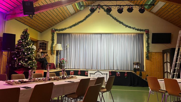 Der Raum ist festlich dekoriert mit einem Weihnachtsbaum und festlichem Tisch. Im Hintergrund steht ein Podium mit einem Vorhang und Beleuchtung.