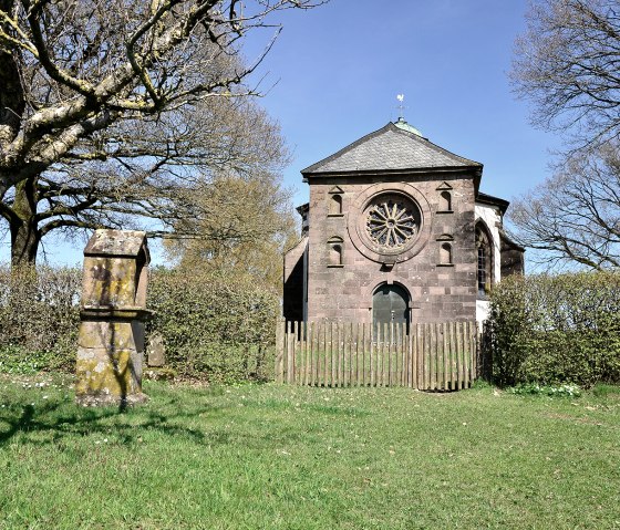 Die Frohnertkapelle in Oberkail, umgeben von Bäumen und einem alten Grabstein im Vordergrund, bei sonnigem Wetter., © TI Bitburger Land