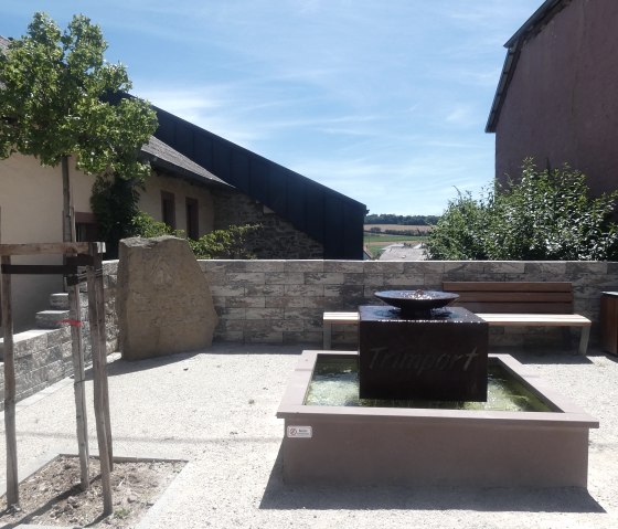Een kleine rustplaats in Trimport met een fontein, een houten bankje en een boom. Op de achtergrond zijn gebouwen en een blauwe lucht te zien., © TI Bitburger Land