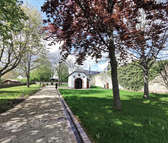 Ein gepflegter Park mit Bäumen, grünem Rasen und einem kleinen weißen Gebäude. Ein gepflasterter Weg führt durch den Park., © Melanie Lonien