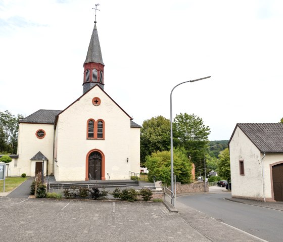 A church in Wißmannsdorf with a pointed tower, surrounded by trees and a parking lot. On the right another building and a road., © TI Bitburger Land