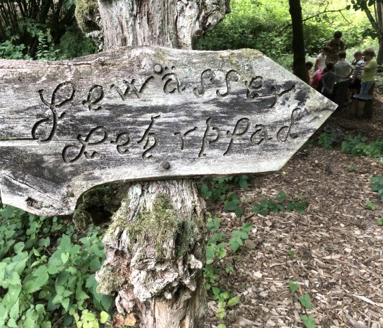 Ein verwittertes Holzschild mit der Aufschrift 'Gewässer Lehrpfad' zeigt den Weg in einen Wald. Im Hintergrund sind Kinder zu sehen., © Tourist-Info Bitburger Land