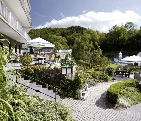 Terrasse des Hotel Haus Kylltal mit Tischen und Sonnenschirmen, umgeben von üppiger Vegetation und Wald im Hintergrund., © Hotel Haus Kylltal