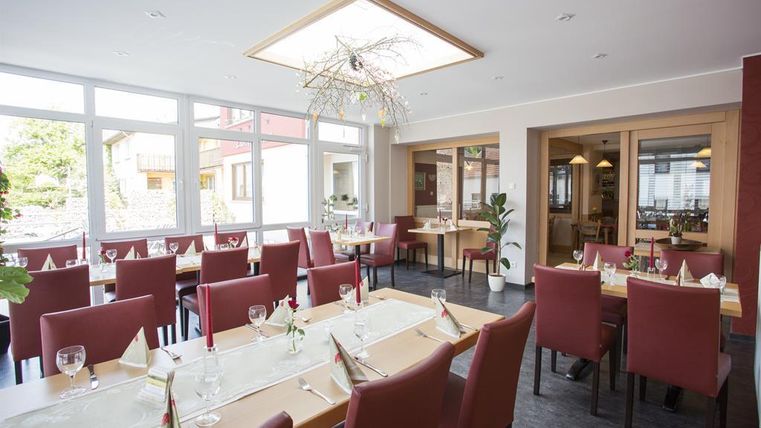 Ein helles Restaurant mit großen Fenstern und eleganten Holztischen. Die roten Stühle und die stilvolle Dekoration schaffen eine einladende Atmosphäre.