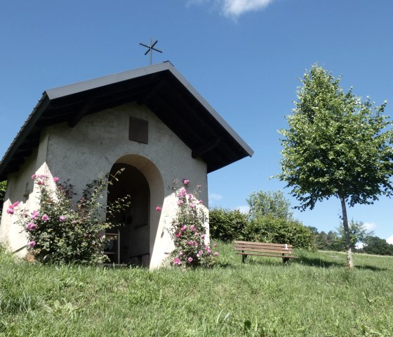 A small chapel in the countryside, surrounded by blooming roses. A bench stands next to it, a tree provides shade. Blue sky in the background., © Tourist-Information Bitburger Land, Melanie Salzburger