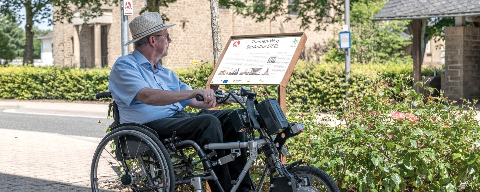 Un homme en fauteuil roulant lit un panneau du sentier thématique Baukultur Eifel à Wolsfeld. En arrière-plan, on aperçoit des arbres et un bâtiment., © Naturpark Südeifel, Thomas Urbany