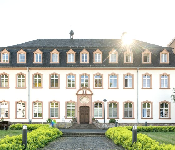 A historic building with a symmetrical façade, lots of windows and a well-tended garden. The sun shines out from behind the roof., © Eifel Tourismus GmbH, Dominik Ketz