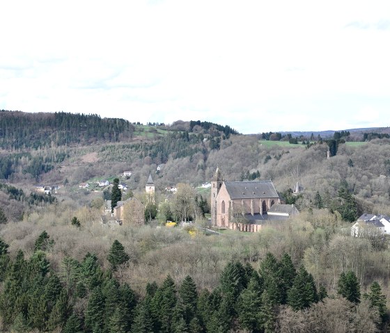 Blick auf eine Kirche inmitten einer bewaldeten Landschaft mit Hügeln im Hintergrund, aufgenommen von der Wilsecker Linde., © TI Bitburger Land