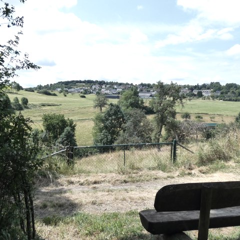 Eine Holzbank steht am Rand eines Feldwegs mit Blick auf eine grüne Landschaft und ein Dorf in der Ferne unter blauem Himmel., © TI Bitburger Land