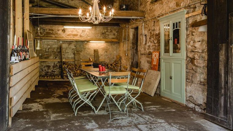 Ein rustikaler Innenraum mit einem langen Tisch und Stühlen. An der Wand befindet sich eine grüne Tür und eine Bar mit Getränken.