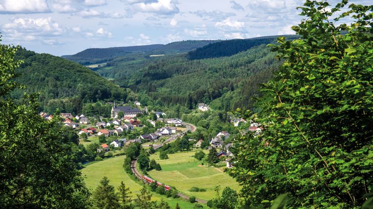 Panoramablick auf ein Dorf in einer grünen, bewaldeten Landschaft mit einem Zug auf einer Bahnstrecke.