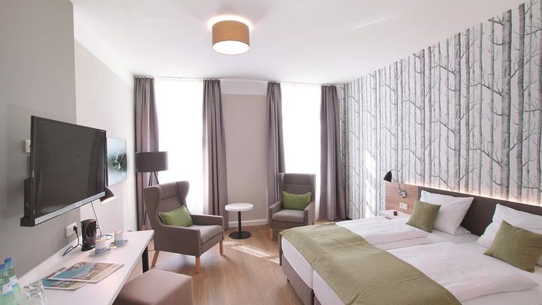 Ein modernes Hotelzimmer mit einem Doppelbett und eleganter Einrichtung. Es gibt Fenster mit Vorhängen, einen Flachbildfernseher und eine gemütliche Sitzecke.