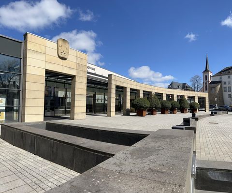 Ein modernes Gebäude mit großen Glasfenstern und einem geschwungenen Design. Im Vordergrund sind Pflanzkästen und ein Platz mit Steinbelag zu sehen.