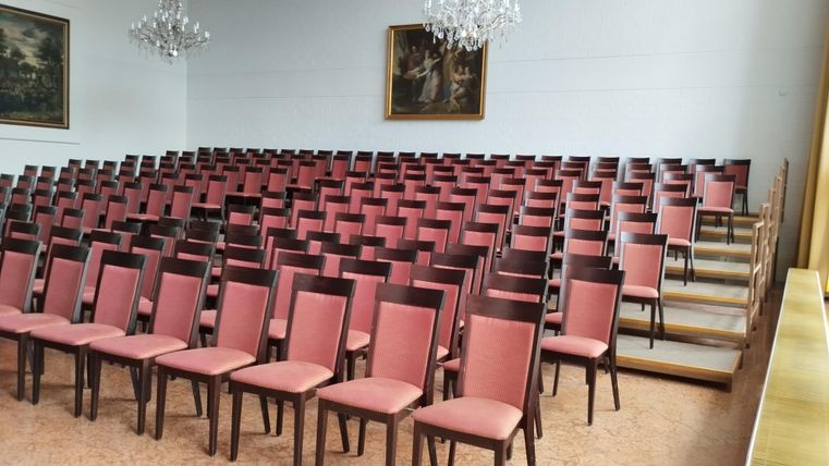 Een grote ruimte met veel rijen roze stoelen. Op de achtergrond zijn kunstwerken en kristallen kroonluchters te zien.