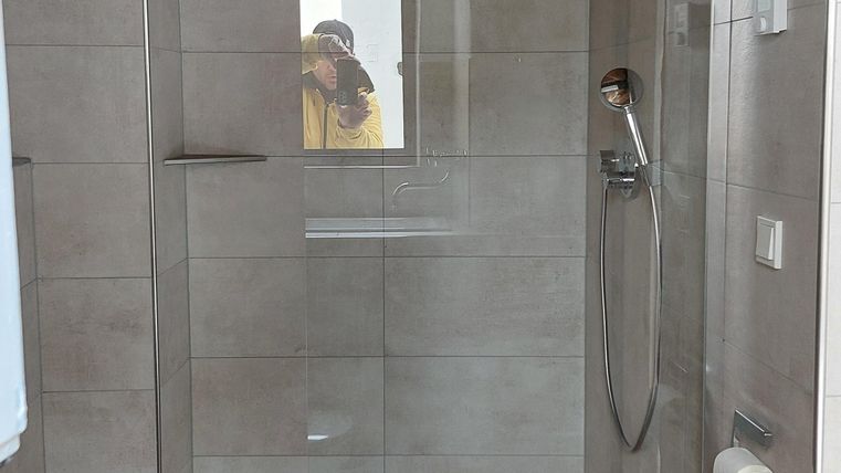 Een moderne douche met glazen scheidingswand en houten plafond. De vloer is betegeld in houtlook.