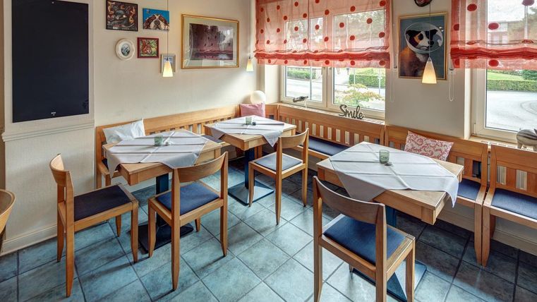 Eine gemütliche Restaurantumgebung mit Holztischen und Stühlen. Rote Vorhänge und dekorative Wandbilder schaffen eine einladende Atmosphäre.