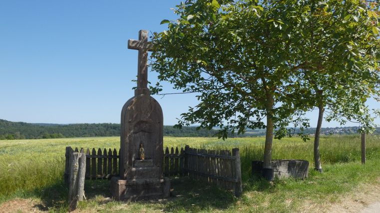 Ein steinernes Wegekreuz steht neben einem Baum auf einem Feldweg, umgeben von einem Holzzaun. Im Hintergrund sind Felder und ein Wald zu sehen.