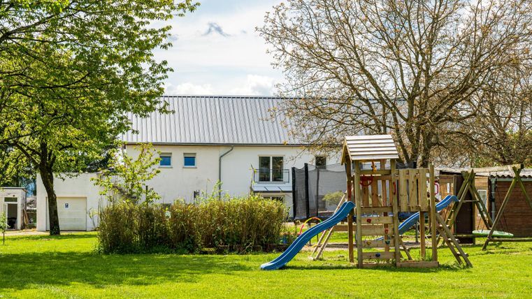 Een speeltuin met een glijbaan en speeltoestel op een groene wei. Op de achtergrond is een helder gebouw en enkele bomen te zien.
