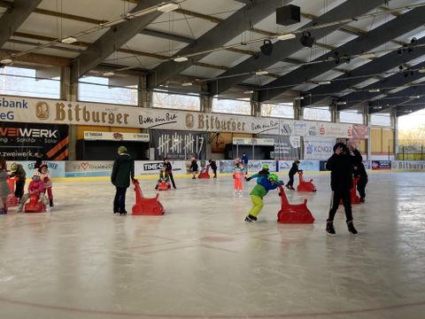 Eine Eishalle mit Kindern, die das Schlittschuhlaufen üben. Einige Kinder nutzen Schlitten zur Unterstützung.