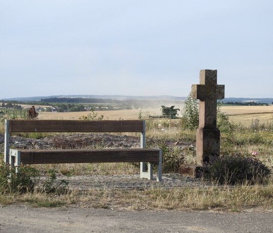 A wooden bench and a stone cross stand on a dirt road. A combine harvester can be seen in a field in the background., © Thomas Neises