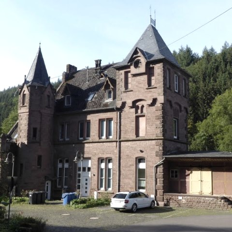 Historisch stationsgebouw van rode baksteen met torens, omringd door bomen en een geparkeerde auto., © Eifelverein Ortsgruppe Speicher