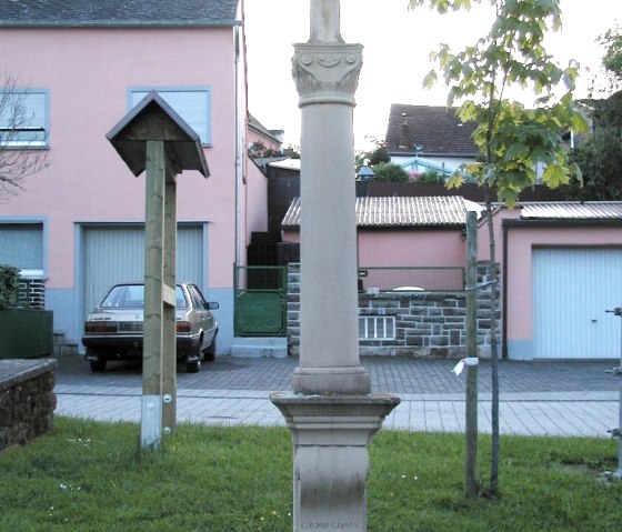 Een stenen wegkruis staat op een dorpsplein, omgeven door gras. Op de achtergrond zie je een boom en een roze huis., © Conny Meier