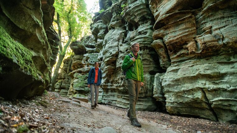 Zwei Wanderer gehen durch eine enge Schlucht mit beeindruckenden Felsformationen. Über ihnen wachsen grüne Bäume, die eine ruhige Atmosphäre schaffen.