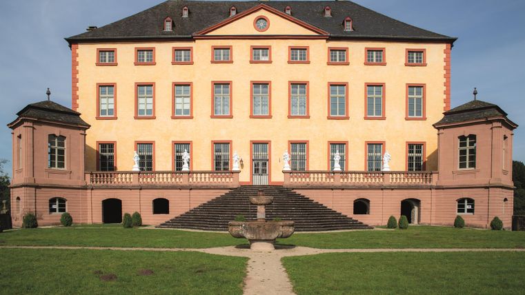 An impressive castle with an orange facade and large windows. It is surrounded by a well-maintained garden and offers a majestic view.