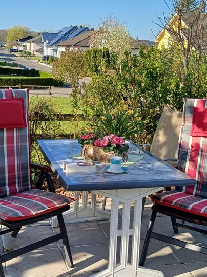 Ein gemütlicher Tisch mit zwei Stühlen im Freien, dekoriert mit Blumen und einer Tasse Kaffee. Im Hintergrund sind grüne Pflanzen und Häuser zu sehen.