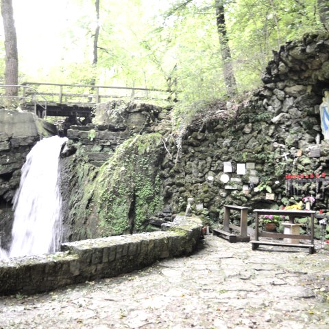 Die Mariengrotte Albach zeigt zwei Wasserfälle, eine Marienstatue in einer Felsnische und Kerzen auf einem Altar. Umgeben von üppigem Grün., © TI Bitburger Land