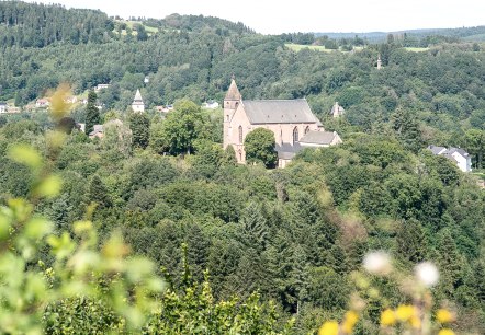 Gezicht op een kerk, omringd door dichte bossen en heuvels, onder een blauwe lucht. Op de voorgrond zijn vage planten te zien., © Tourist-Info Bitburger Land M.Mayer