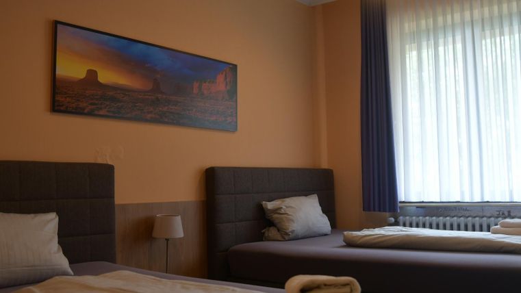 Ein einfaches Zimmer mit zwei Betten und einem Gemälde an der Wand. Es gibt große Fenster, die viel Tageslicht hereinlassen.